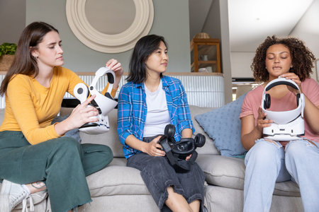 Diverse female friends adjusting white VR headsets and holding controllers on sofa in living room....
