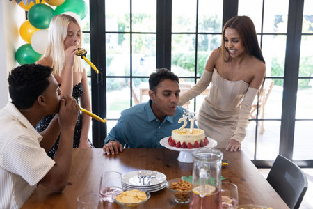 Diverse friends celebrating 21st birthday in home dining room with cake, balloons, party blowers....