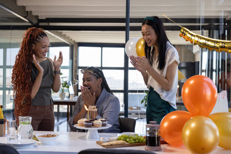 Diverse female coworkers celebrating birthday around office table with lit cupcakes and gold banner....