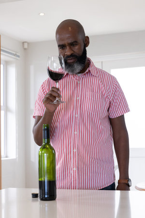 Senior African American man examining red wine in stemmed glass at modern kitchen counter. Luxury,...