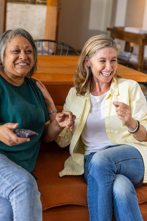 Senior female friends chatting animatedly on rust-colored sofa in living room with TV remote....