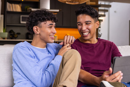 Two male friends laughing and sharing tablet on light-colored sofa in modern living room. Casual,...