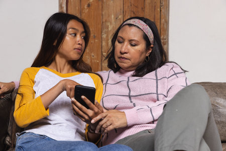 Diverse mother and daughter sitting on sofa in living room scrolling smartphone next to wooden door....