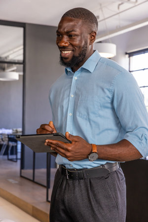 African American man wearing shirt holding tablet and stylus while reviewing plans in office space....