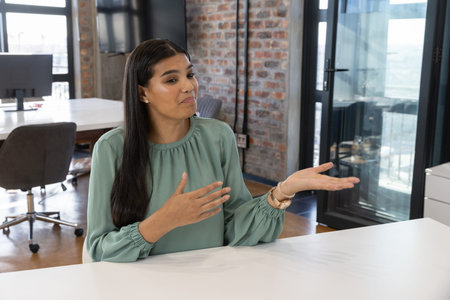 Woman wearing green blouse sitting at conference table in office gesturing and speaking by monitor....