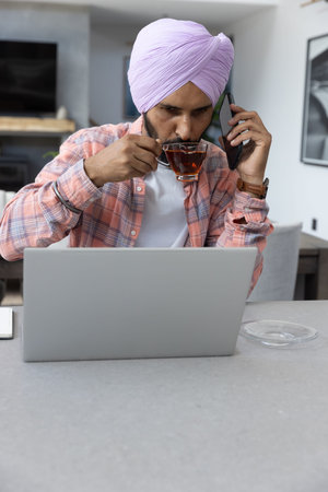 Man wearing lavender turban using smartphone and sipping tea at countertop with open laptop....