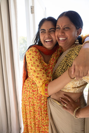 Indian mother and daughter hugging and laughing by sliding glass door wearing saree and kurta....