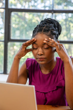 African American woman pressing temples, closing eyes at wooden desk with laptop by window. Stress,...