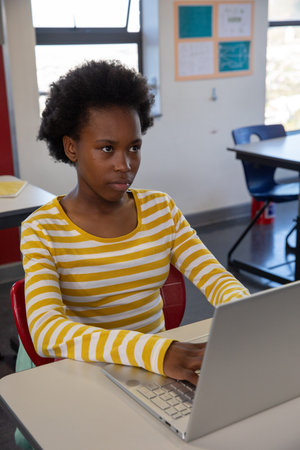 African American teen girl typing on silver laptop at white desk in classroom wearing striped shirt....
