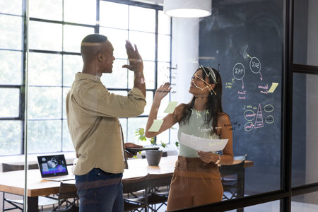 Diverse coworkers giving high-five while writing sticky notes on glass whiteboard in meeting room....