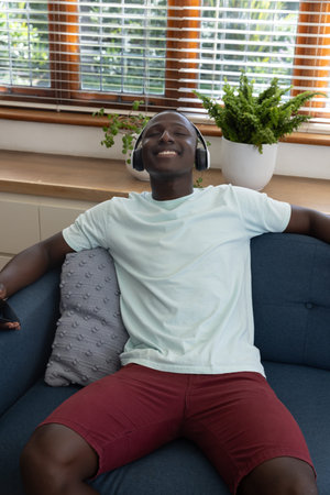 African American man reclining on sofa wearing wireless headphones beside plant pots on windowsill. Relaxation, comfort, leisure, modern, minimalist, tranquility, homeの写真素材