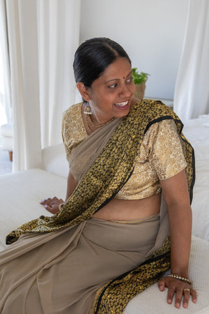Mature Indian woman sitting on edge of bed in bedroom near plant wearing saree and jewelry. Elegant,...