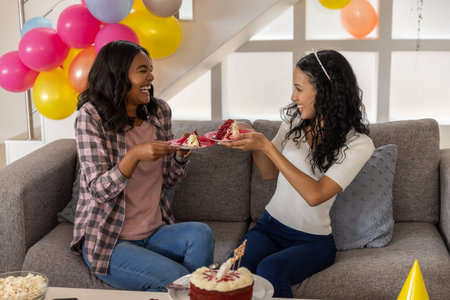 Diverse female friends sharing cake slices on couch in living room with balloons and birthday cake....