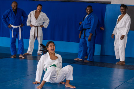 Teenage girl in gi and green belt laughing on mat at dojo with partners observing. Martialarts,...