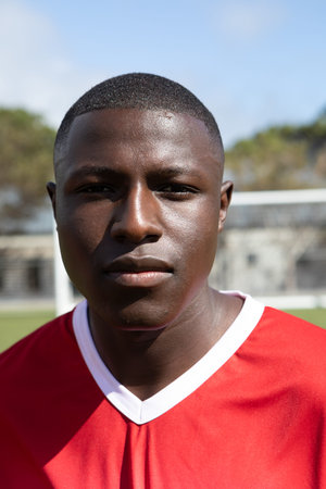 African American male standing on soccer field wearing red jersey with white collar and goal net....