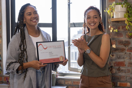 Diverse coworkers celebrating certificate at office window, clapping near potted plant and mug....