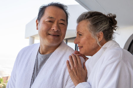 Diverse senior couple standing on spa balcony, wearing white bathrobes and gazing at each other....
