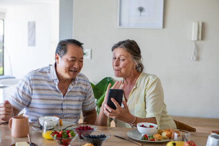 Diverse couple sitting at home dining table smiling at smartphone with fresh berries and croissants....