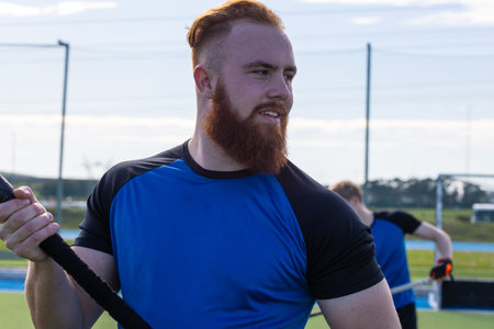 Male teammates wearing blue-black sportswear gripping braided rope and pulling on artificial turf....