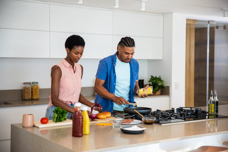 Diverse couple slicing onion and lettuce while sprinkling cheese on frying pan at kitchen island....