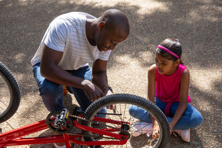 Diverse father and daughter examining red bicycle front wheel on paved park path. Adventure, outdoor, bonding, family, exploration, vitality, recreationの写真素材