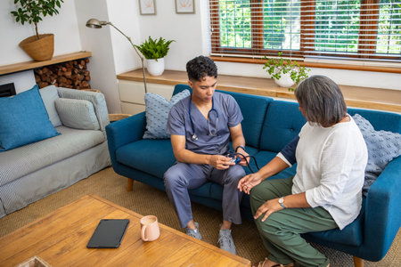 Diverse senior patient and caregiver sitting on sofa checking blood pressure with stethoscope....