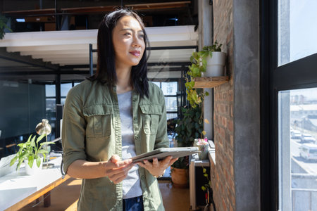 Asian woman standing in bright office space by large window holding tablet and surveying city view....