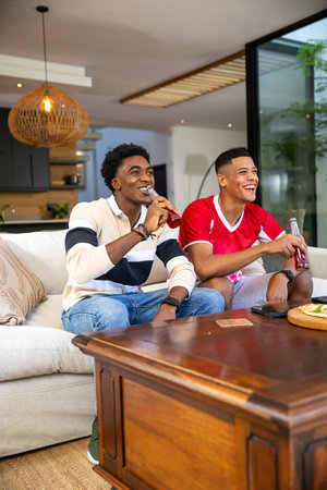 Diverse male friends smiling and holding glass bottles on sofa near coffee table with snack platter....