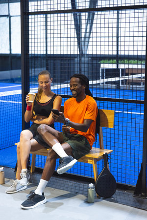 Diverse friends in sportswear sitting on padel court bench holding coffee cup smartphone and racket....