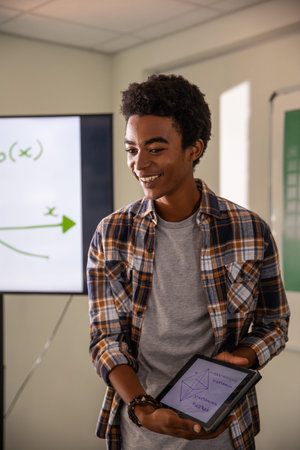 African American male holding tablet with diagrams in class, monitor showing graph and whiteboard....