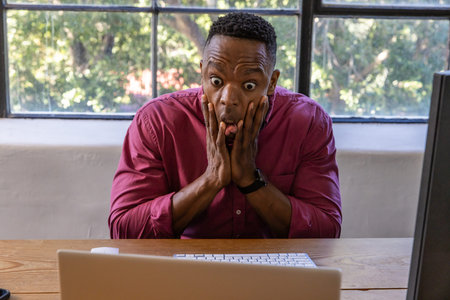 African American man pressing cheeks and showing shock at home desk with laptop and smartwatch....
