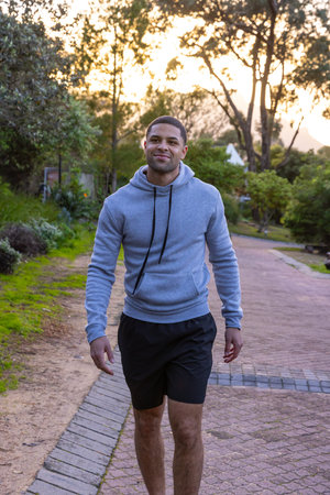 African American male walking brick-paved path through verdant park at sunset in hoodie and shorts....