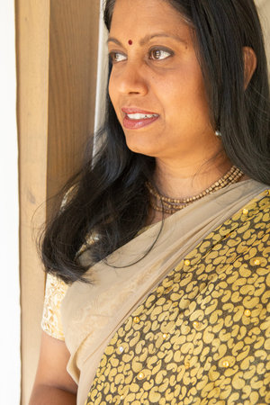 Indian woman leaning against wooden window frame inside home with cream sari and bead necklaces....