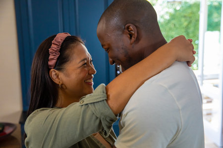 Diverse couple hugging and gazing into each other's eyes in home kitchen with pink headband....