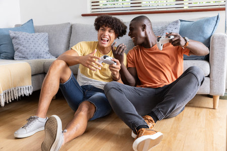 Diverse friends laughing with game controllers while sitting on wooden floor before gray sofa....