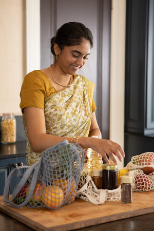 Indian woman in floral sari arranging glass jars and mesh produce bags on wooden kitchen island....
