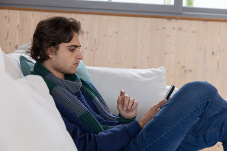 Man sitting on sofa with cushion in living room wearing scarf holding smartphone. Casual,...