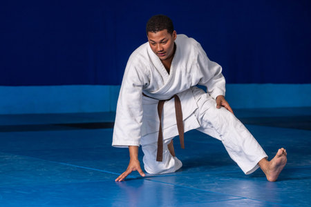Adult male martial artist kneeling, pressing on blue dojo mat in white gi and brown belt. Martial...