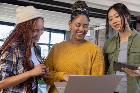 Diverse female coworkers reviewing content on laptop at modern office desk with tablet....