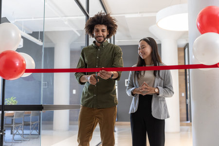 Diverse coworkers cutting red ribbon at office lobby with glass partitions, balloons and scissors. Celebration, professionalism, milestone, ribbon-cutting, cooperation, architecture, contemporary