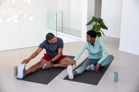 African American fitness partners stretching hamstrings on mats in bright gym with water bottles....
