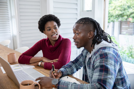 African American colleagues collaborating over laptop and notebook with mugs at home. Collaboration,...