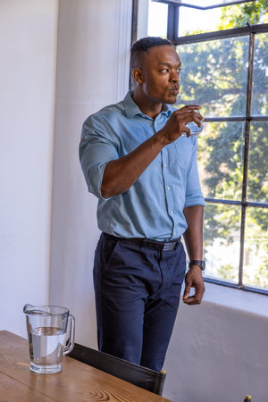 African American man in 30s standing beside window in office holding tumbler near pitcher on table....