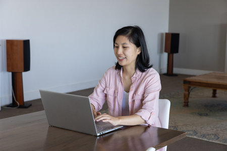Asian woman sitting at wooden desk working on silver laptop in home office with speakers....