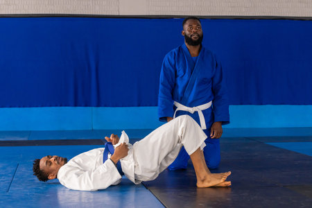 African American male training partners practicing hip bridge in gym wearing gis and belts on mats....