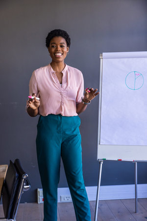 African American woman holding colored markers and presenting beside flipchart near table and chair....