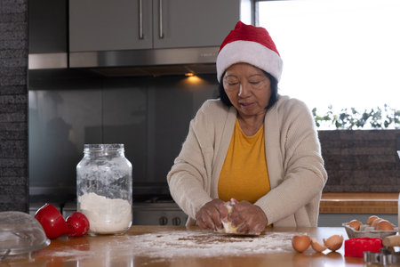 Senior Asian woman wearing red Santa hat kneading dough on kitchen countertop with flour jar....