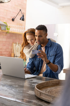 Diverse couple hugging while using laptop and smartphone on countertop table with wicker tray. Contemporary, cozy, lifestyle, interior, warmth, affection, communicationの写真素材