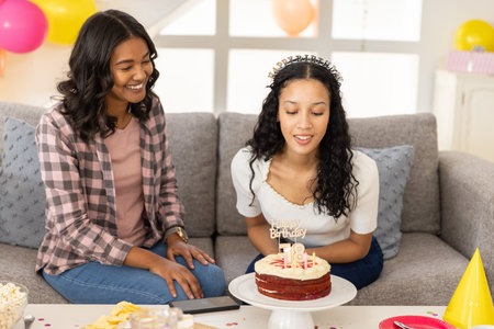 Diverse female friends celebrating eighteenth birthday on grey couch at home with cake and balloons....