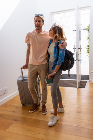 Couple standing in bright entryway holding rolling suitcase and passport while wearing backpack....